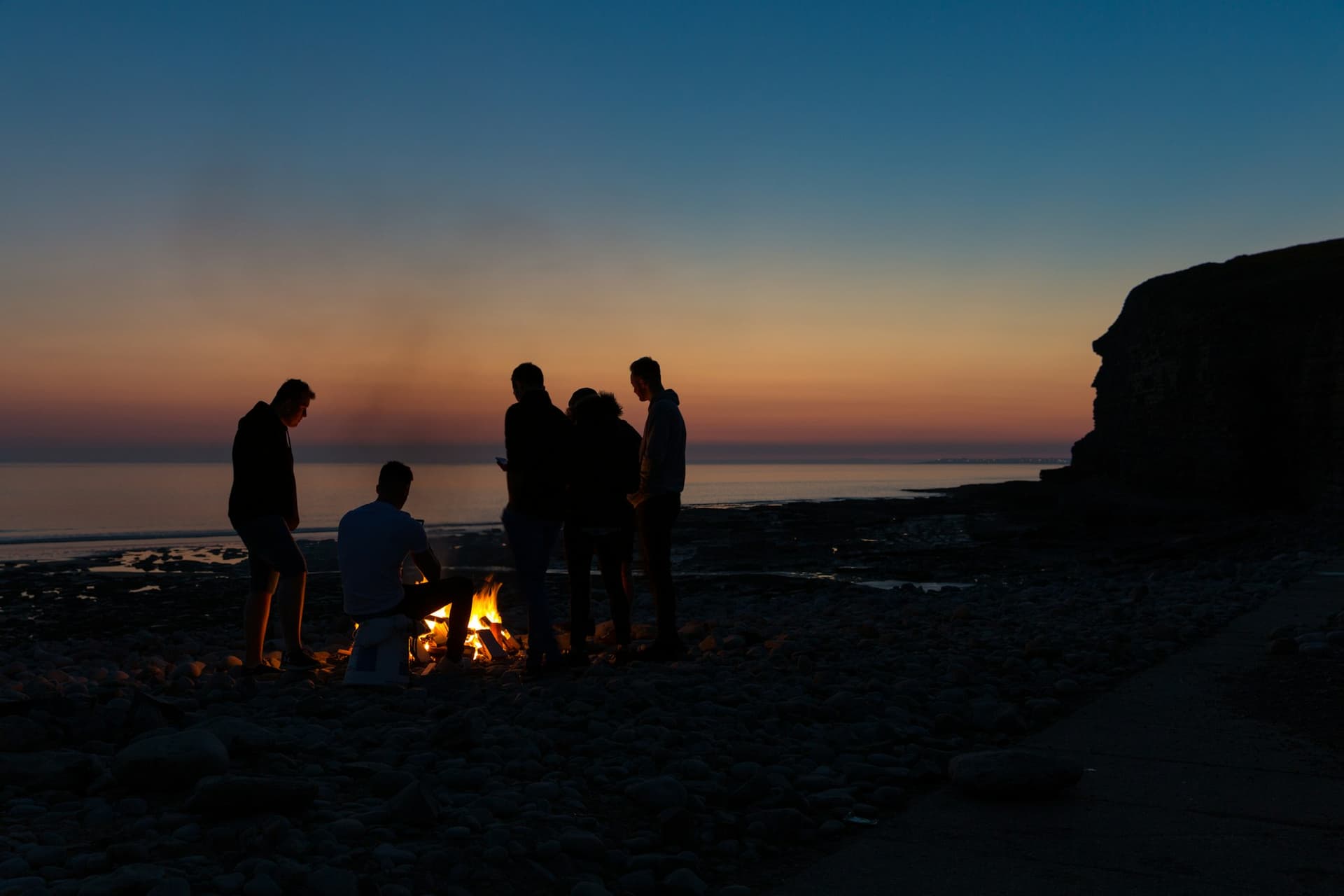 Friends in silhouette gathered around a small campfire on a pebble beach after sunset