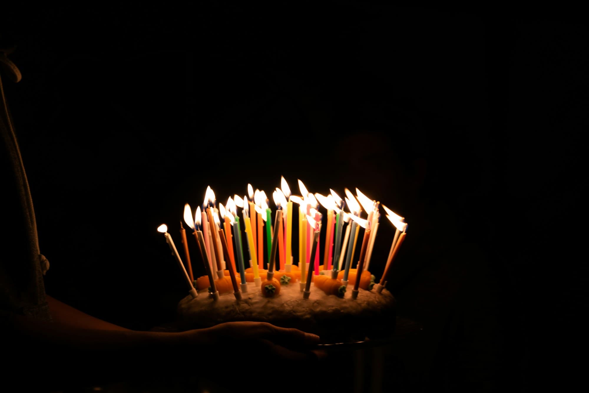 Hands carrying a birthday cake crowded with lit candles through a dark room